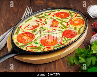 Poêle en fonte ovale avec délicieux chaud omelette aux tomates et poivrons. Brown wood texture background, close-up, vue de côté. Banque D'Images