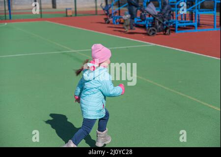 Un enfant dans une veste bleue court autour d'un terrain de sport vert. Parents et grand-mère avec des voitures d'enfant en arrière-plan. Un homme espiègle de cinq ans Banque D'Images