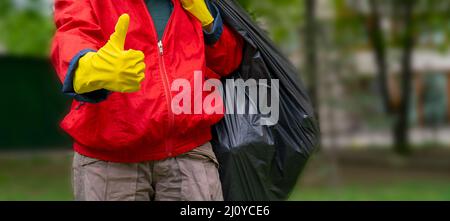 La collecte des déchets. L'homme en manteau rouge et jaune gants showing thumb up, porte un gros sac noir avec corbeille. Banque D'Images