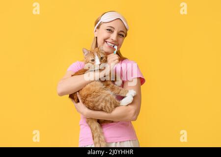 Belle femme mûre avec chat mignon se brossant les dents sur fond jaune Banque D'Images
