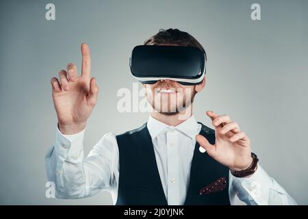 Immergée dans un environnement d'entreprise virtuel. Photo en studio d'un jeune homme d'affaires charmant utilisant un casque vr sur fond gris. Banque D'Images