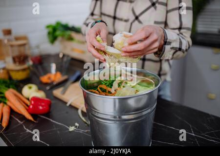 Femme jetant des boutures de légumes dans un seau de compost dans la cuisine. Banque D'Images