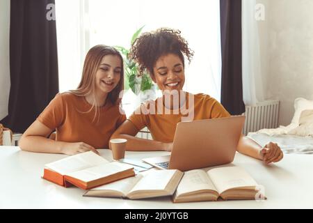 Deux amies heureuses assises devant un ordinateur portable, préparant les examens ensemble à la maison. Banque D'Images