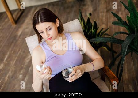 Suppléments pendant la grossesse. Jeune femme enceinte heureuse prenant des vitamines prénatales, tenant un verre d'eau et de pilule tout en étant assise à un fauteuil à la maison. Banque D'Images