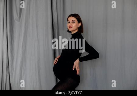 Jeune femme enceinte debout sur fond gris regardant l'appareil photo touchant le ventre en portant des vêtements noirs Banque D'Images