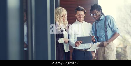 Faire des affaires sur le balcon. Prise de vue courte de trois jeunes hommes d'affaires utilisant une tablette tout en se tenant à l'extérieur sur le balcon du bureau. Banque D'Images