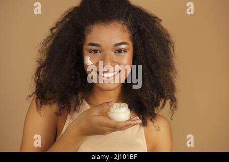 Gros plan d'une femme à la peau sombre et souriante, avec des cheveux freux qui appliquent une crème pour le visage Banque D'Images