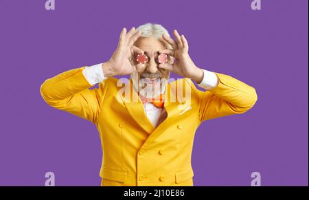 Portrait de drôle heureux senior homme en costume jaune couvrant ses yeux avec deux jetons de poker Banque D'Images