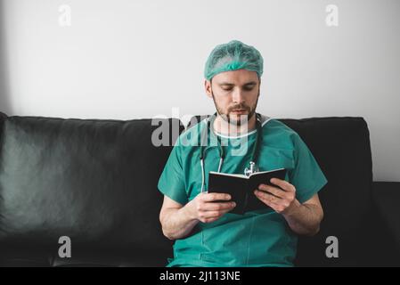 Médecin lisant un livre dans la salle de repos, portant une robe chirurgicale Banque D'Images