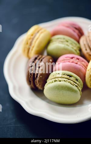 Macarons français à la framboise, au pistache, au chocolat et au citron sur une assiette blanche vintage avec un fond noir Banque D'Images