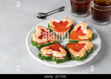 Canape sous forme de coeurs avec caviar rouge et crevettes sur une plaque blanche sur fond gris clair, idée de petit déjeuner de la Saint-Valentin Banque D'Images