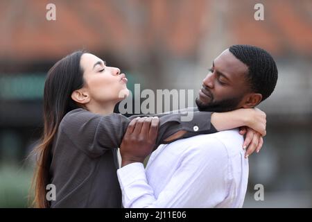 Vue latérale portrait d'une femme obsédée essayant d'embrasser un homme à la peau noire qui la rejette dans la rue Banque D'Images