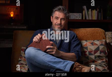 Portrait d'un homme plus âgé à la maison dans une pièce sombre, assis sur un canapé. Sourire heureux, cheveux gris, barbu, . âge mûr, âge moyen, homme adulte moyen en 50s. Banque D'Images