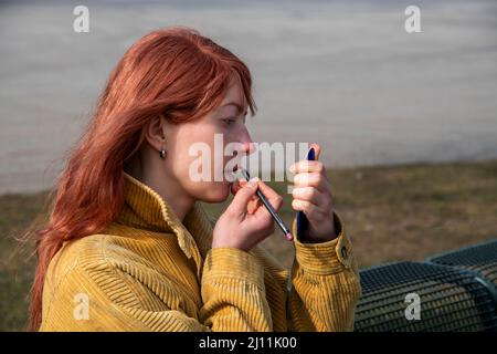 Vue latérale d'une femme aux cheveux rouges qui se fait, colorant ses lèvres Banque D'Images