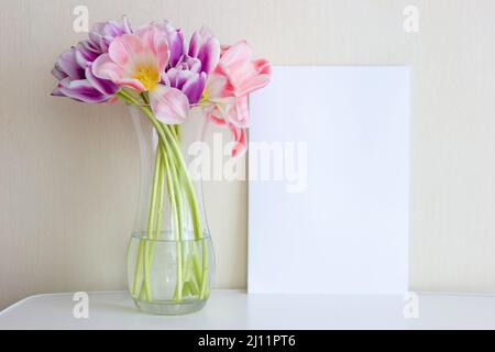 Un bouquet de tulipes roses ouvertes dans un vase en verre se dresse sur une table blanche contre un mur léger. Banque D'Images