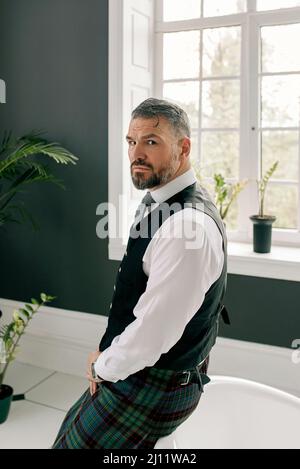 beau mûr courageux et élégant homme d'affaires écossais dans le kilt et le costume dans la salle de bains. Style, travail à la maison, mode, style de vie, culture, ethnique Banque D'Images