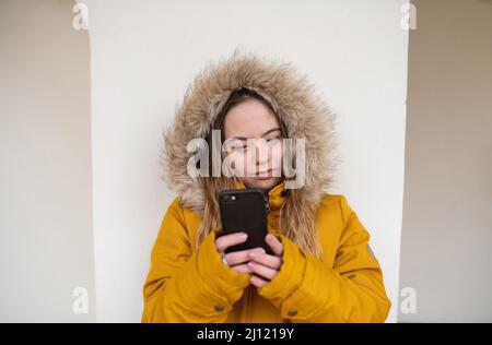 Jeune femme heureuse avec le syndrome de Down weraing parka, se penchant le mur et en utilisant le smartphone. Banque D'Images