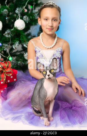 Sphynx roux regardant l'appareil photo, assis près de neuf ans enfant en robe mauve sur le fond de l'arbre de Noël avec des boîtes-cadeaux à pois rouges Banque D'Images