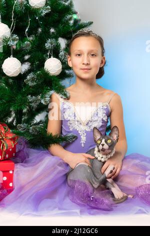 Fille en robe violette tenant le chat sans poils Sphynx purébred se trouve près de l'arbre de Noël avec des boîtes-cadeaux rouges à la Saint-Sylvestre. Mignon neuf ans Banque D'Images