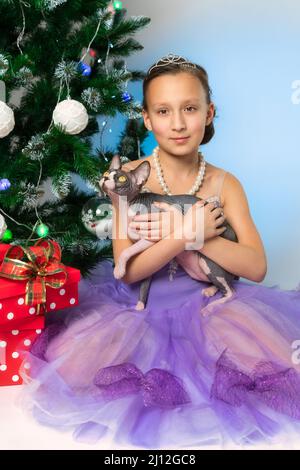 Fille en robe violette tenant le chat sans cheveux Sphynx espiègle se trouve près de l'arbre de Noël avec des boîtes cadeau à pois rouges à la Saint-Sylvestre. Enfant pré-adolescent Banque D'Images