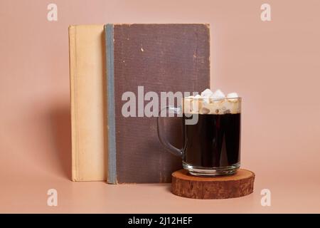 Une tasse de café en verre avec guimauves se tient sur un support en bois à côté des livres Banque D'Images