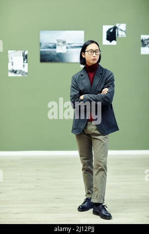 Prise de vue verticale en longueur d'un jeune asiatique élégant portant des lunettes debout avec des bras croisés dans une galerie d'art moderne Banque D'Images