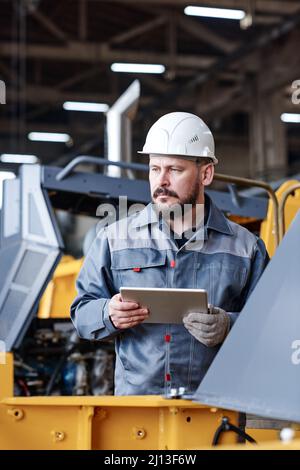 Travailleur masculin sérieux et mature du service de gestion du contrôle de la qualité utilisant une tablette lors du test de nouvelles machines industrielles Banque D'Images