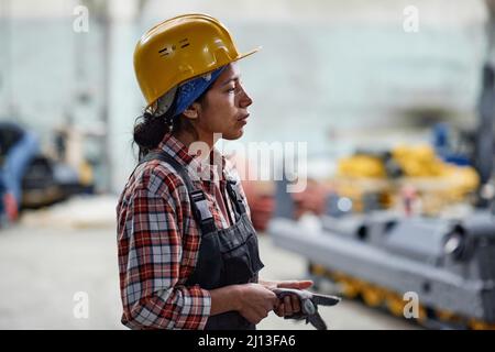 Vue latérale d'une jeune femme fatiguée de l'usine dans un casque et une combinaison debout devant la caméra dans un grand entrepôt Banque D'Images