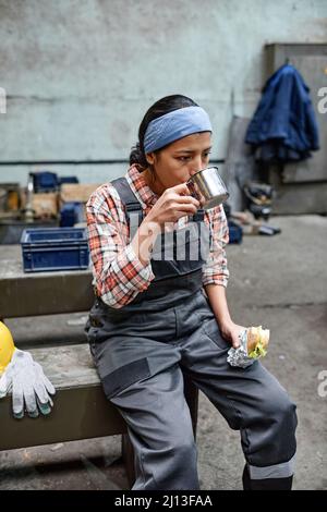 Jeune femme fatiguée et affamée ingénieur en combinaison prenant du thé avec un sandwich pour déjeuner après le travail dans l'entrepôt Banque D'Images