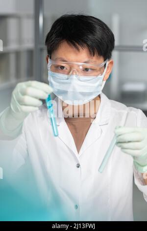 Jeune scientifique asiatique d'origine ethnique tenant une fiole avec le liquide bleu en main et la regardant pendant la recherche scientifique Banque D'Images