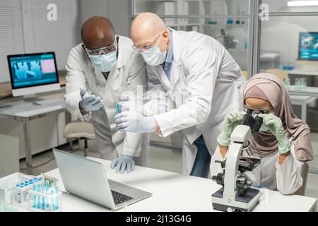 Des scientifiques interraciaux discutent du liquide bleu dans une fiole à côté d'une jeune virologiste musulmane féminine étudiant le nouveau virus au microscope Banque D'Images