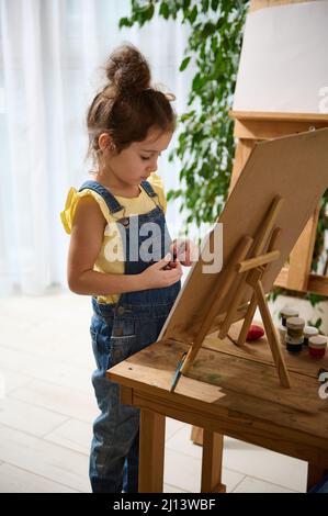 Magnifique peintre caucasien de fille de bébé, petit artiste en t-shirt jaune et bleu denim se tient sur un chevalet en bois à la maison, appréciant la peinture jour créateur Banque D'Images