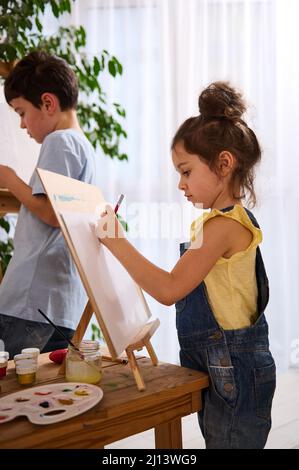 Portrait latéral d'une petite fille heureuse en t-shirt jaune et bleu denim dessin image avec son frère. Peintures à l'huile et à l'aquarelle avec des brosses sur un bon de Banque D'Images