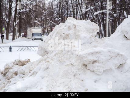 Une grande dérive de neige au bord de la route sur fond de rue de la ville. Sur la route se trouve de la neige sale dans des tas hauts. Paysage urbain d'hiver. Jour d'hiver nuageux, lumière douce. Banque D'Images