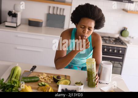 Femme afro-américaine de taille moyenne adulte qui se léchant le doigt tout en buvant du jus sain dans la cuisine à la maison Banque D'Images