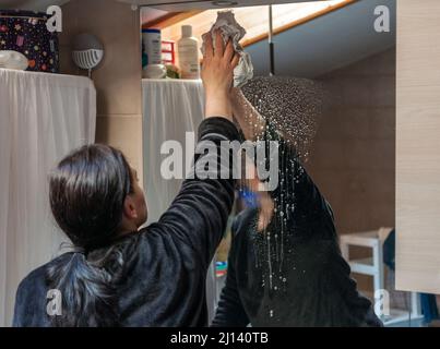 femme de ménage essuie un miroir avec un chiffon. vu de l'arrière et partiellement reflété dans le miroir Banque D'Images