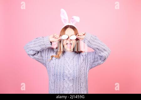 Belle fille dans un chandail bleu sur fond rose. Femme aux oreilles de lapin, posant pour pâques, couvrant ses yeux avec des œufs de pâques Banque D'Images