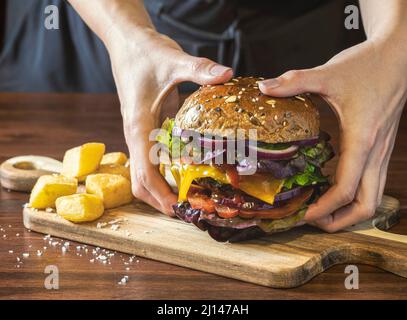 Gros plan d'un délicieux hamburger avec des légumes et des frites sur une table en bois sombre. Une personne méconnaissable prend le hamburger avec deux mains pour le manger. Banque D'Images