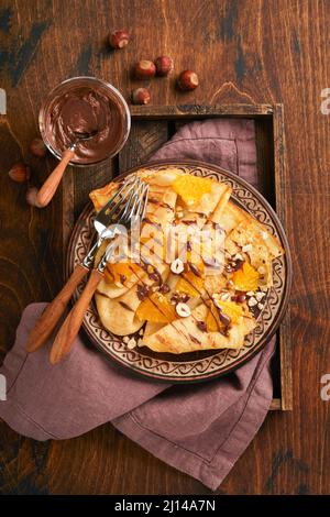 Crêpes suzette aux oranges et au chocolat. Fines crêpes avec tartinade au chocolat, noisettes et tranches d'orange dans l'assiette pour le petit déjeuner sur le vieux woo sombre Banque D'Images