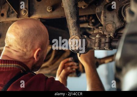 Le mécanicien ou mécanicien de voiture à chauve est assis sous la machinerie et répare la suspension du moteur de la voiture dans l'atelier. Entretien des camions lourds. Vue de l'arrière. Service de voiture. Arrière-plan. Banque D'Images