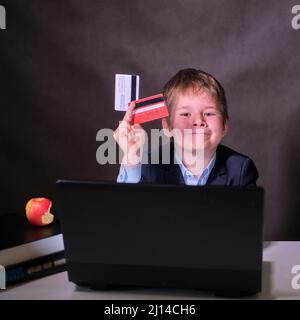 Un garçon souriant dans un costume d'école avec des cartes de crédit à l'ordinateur, un espace de copie sur un arrière-plan sombre de studio Banque D'Images