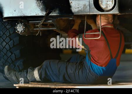 Le serrurier ou le mécanicien de voiture est assis sur le sol sous la voiture et répare les gros équipements dans le garage. Service de voiture. Arrière-plan. Banque D'Images