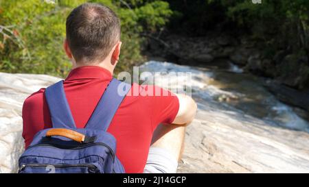 Homme voyageur dans un polo rouge avec un sac à dos bleu se trouve près d'une rivière de montagne Banque D'Images