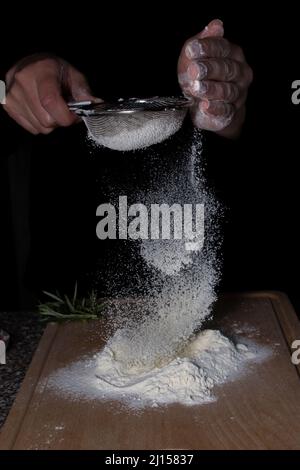 Femme chef mains tenant un tamis avec de la farine tombant sur le bois avec du romarin sur la table. Banque D'Images
