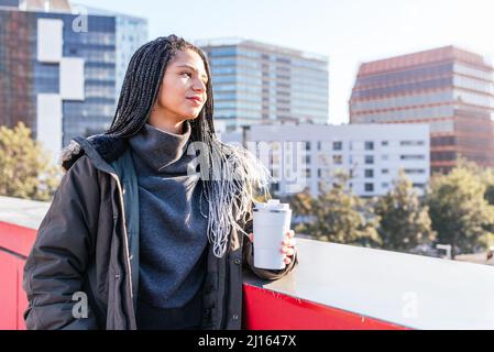 Jolie femme hispanique en vêtement d'extérieur chaud avec thermos de café chaud debout près de la frontière tout en observant la ville avec des bâtiments résidentiels Banque D'Images