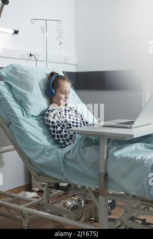 Petite fille malade portant un casque et regardant des dessins animés sur un ordinateur portable tout en se reposant dans la salle de clinique pédiatrique. Pas bien petite fille utilisant un ordinateur portable pour regarder des vidéos drôles tout en étant assis dans le lit d'hôpital. Banque D'Images