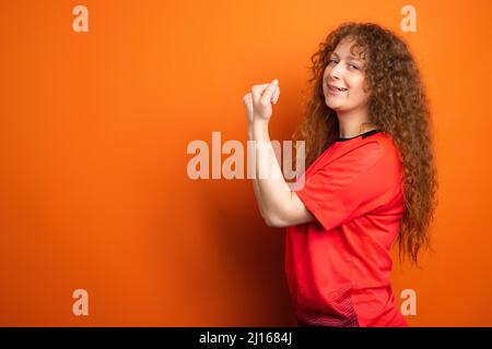 La jeune femme en uniforme de football qui garde des poings pour son équipe de football préférée comme gagnante isolée au studio orange. Banque D'Images