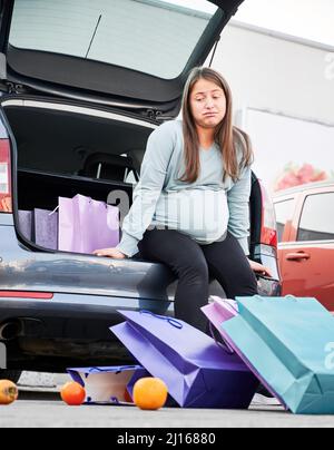 Vue de dessous de la femme enceinte contrariée assise dans le coffre ouvert de la voiture, regardant les sacs de couleurs éparpillées des oranges avec l'expression mécontente sur son visage. Banque D'Images