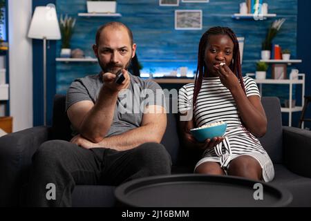 Homme caucasien et femme afro-américaine regardant la télévision tout en partageant un bol de pop-corn assis sur le canapé à la maison. Mari stressé changer de programmes de télévision à la recherche d'un film. Banque D'Images