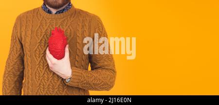 Jeune homme caucasien barbu tenant le coeur artificiel dans sa main, l'accent est sur le coeur, aucun visage visible, pull maladie cardiaque concept isolé jaune fond studio tourné . Photo de haute qualité Banque D'Images